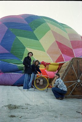 Vibrant 1970s-style hot air balloon with geometric, rainbow-colored panels being inflated by three people on sandy ground. Tw...