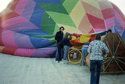 Vibrant geometric hot air balloon in mid-inflation, featuring bold pink, green, and purple panels. Four people in 1970s-style...