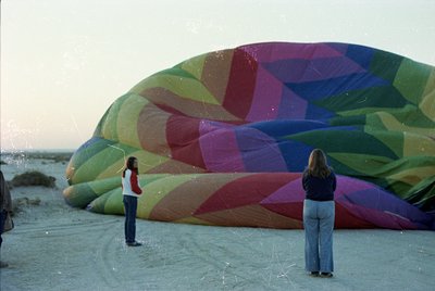 Two individuals stand on a wet beach, inspecting a large, geometric-patterned hot air balloon with vibrant stripes in green, ...