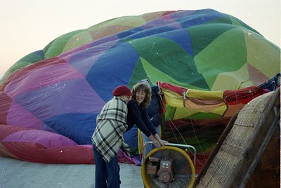 Vibrant 1970s hot air balloon in deflated state, with two people preparing for launch. The balloon’s fabric features bold geo...
