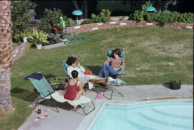 Vintage backyard pool scene with three adults lounging on green metal chairs, 1970s-80s suburban style. Concrete pool with sh...