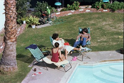 Aerial view of a mid-20th-century backyard pool scene. Three adults relax on vintage lounge chairs beside a rectangular pool,...