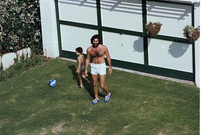 Vintage 1970s backyard scene: shirtless man in white shorts and blue sneakers plays with toddler in matching swimwear. Grass,...