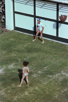 Vintage 1960s backyard scene: two children playing barefoot on a grassy lawn, one in a striped swimsuit, the other in shorts ...