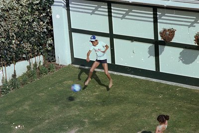 Mid-20th century suburban backyard scene: boy in white sports shirt and blue cap kicks a blue ball toward a garage door. Lush...