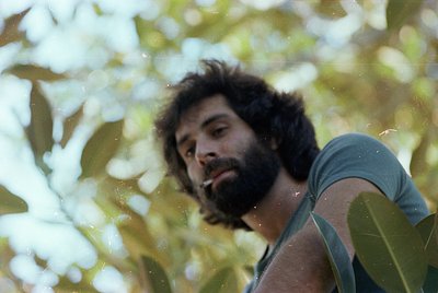Portrait of a bearded man in a sleeveless shirt, leaning against lush green foliage. His relaxed pose suggests a candid, outd...