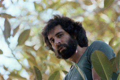 Portrait of a man with a full beard and shoulder-length hair, partially obscured by lush green foliage. The vintage aesthetic...