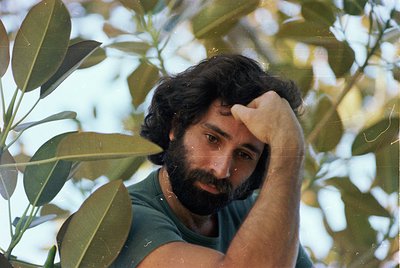 Man in olive grove, hand resting on forehead, mid-20th century attire ( ). Lush green foliage frames his face, suggesting Med...