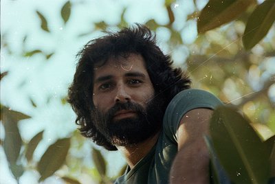 Portrait of a bearded man in a natural setting, framed by lush green foliage. His long hair and mustache suggest a mid-20th-c...