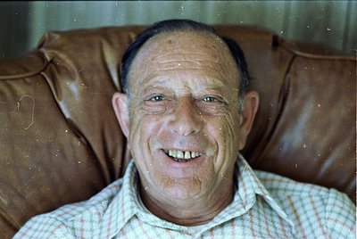 Portrait of an elderly man in a vintage checkered shirt, seated on a worn leather couch. Soft focus and slight film grain sug...