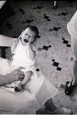 Vintage black-and-white photo of an infant crying while being held by an adult in a clinical setting, likely a hospital or cl...
