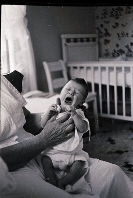 Mid-20th century indoor scene: elderly woman holding crying infant in a simple bedroom. Wooden crib with floral wallpaper in ...