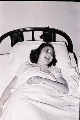 Mid-20th century hospital bed with metal frame, featuring a relaxed woman in a light hospital gown. Classic 1950s–1960s medic...