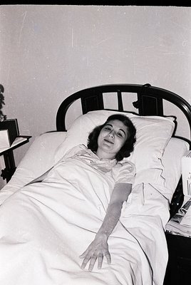 Mid-century hospital bed with metal headboard, featuring a woman in a loose blouse and high-waisted skirt, resting on white l...