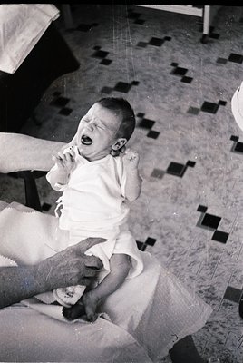 Mid-20th century black-and-white photo of an infant crying in a highchair, wearing a diaper and short-sleeve onesie. Checkere...