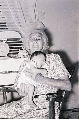 Mid-20th century black-and-white portrait of an elderly woman cradling a baby in a floral dress, seated on a wooden chair. Ba...