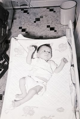 Vintage black-and-white photo of an infant lying on patterned bedding in a crib, mid-20th century. Checkered floor tiles and ...