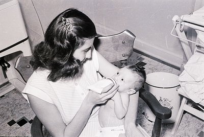 Nurse in 1950s-60s uniform bottle-feeding an infant in a clinical setting, likely a hospital or clinic. Metal feeding chair a...