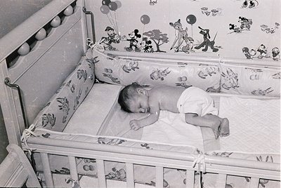 Mid-20th century baby crib with Mickey Mouse-themed decor, featuring cartoon characters and balloons. Infant sleeps peacefull...