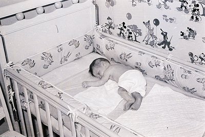 Vintage black-and-white photo of an infant sleeping in a crib adorned with 1960s cartoon-themed bedding (Mickey Mouse, Donald...