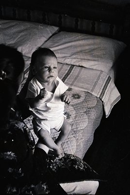 Vintage black-and-white photo of an infant seated on a patterned blanket, likely mid-20th century. The child wears a short-sl...