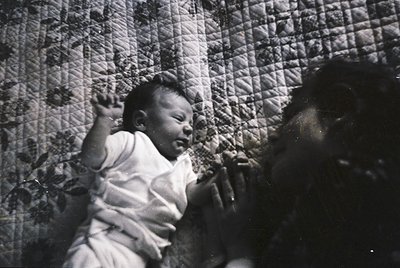 Vintage sepia-toned photo of an infant in a quilted crib, mid-20th century. The geometric patterned fabric suggests mid-centu...