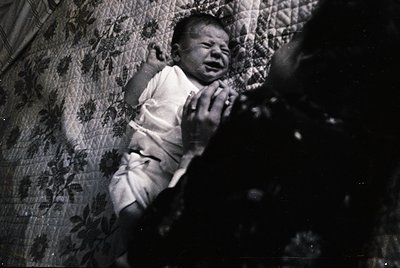 Black-and-white portrait of an infant crying in a crib, draped in floral-patterned bedding. Mid-20th century domestic interio...