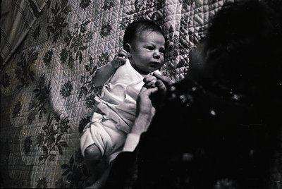 Vintage black-and-white photo of an infant in a diaper, suspended mid-air by an unseen adult’s hands. Floral-patterned wallpa...