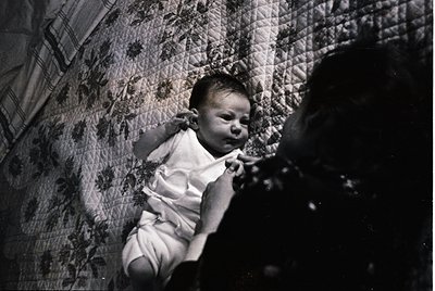 Black-and-white candid of an infant in a patterned quilted bed, mid-cry, with a blurred adult’s hand offering comfort. Mid-20...