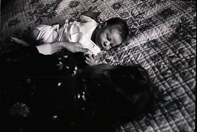 Mid-20th century black-and-white photo of an infant in a simple white dress, lying on a textured quilted blanket. The composi...