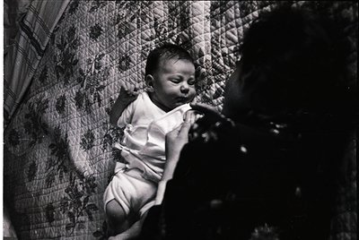 Black-and-white portrait of an infant in a hammock-style bed, draped in a woven, textured fabric. The child wears a simple sh...