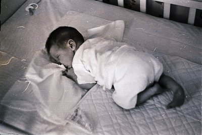 Vintage black-and-white photo of an infant sleeping on a flat mattress in a crib, wearing a long-sleeve onesie. The image ref...