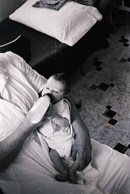 A tender black-and-white moment: an infant in a diaper sits on a bed, being gently held by an adult’s arm. The child’s hand r...