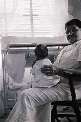 Mid-20th century hospital scene: elderly man cradles newborn in white diaper, seated on a wooden chair beside an iron crib. S...