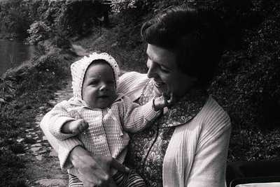 A mid-20th-century black-and-white photo of a woman cradling an infant outdoors near a rocky stream. The woman wears a light ...