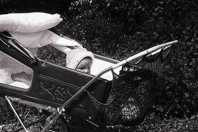 Vintage black-and-white photo of a child in a vintage pram with ornate metalwork, resting on grass. A fishing net and rod lie...