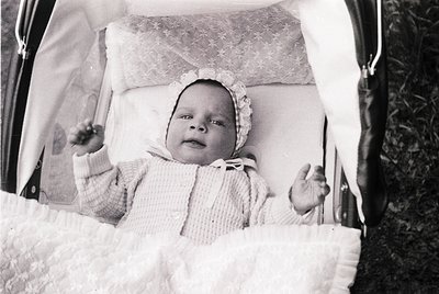 Vintage black-and-white photo of an infant seated in a vintage stroller, dressed in a knitted onesie and bonnet. Soft focus a...