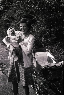 A woman in 1960s-era patterned dress and cardigan holds a baby in a striped onesie, posing outdoors near a vintage pram. Lush...
