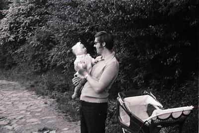 Mid-20th century black-and-white photo of a man in a sweater and glasses holding a baby outdoors, with a vintage pram in the ...