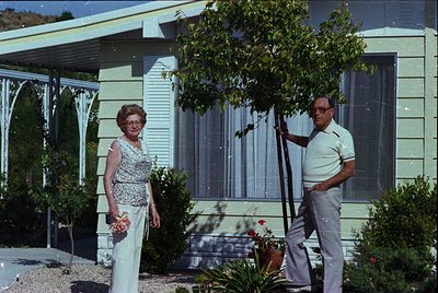 Mid-century modern home exterior with curved roof and sliding glass doors, 1960s USA. Elderly couple in retro attire—woman in...