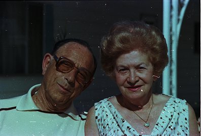 Vintage portrait of an elderly couple posing indoors, likely mid-20th century. Man wears round glasses and a white polo shirt...