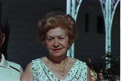 Vintage portrait of an elderly woman in a floral blouse, 1960s-70s style. Soft focus and slight color shift suggest aged film...