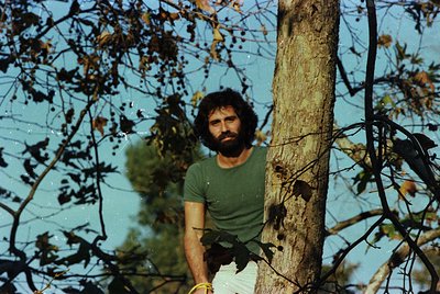 Man in 1970s-style casual wear leans against a tree trunk, partially obscured by autumn foliage. Vibrant green shirt and ligh...