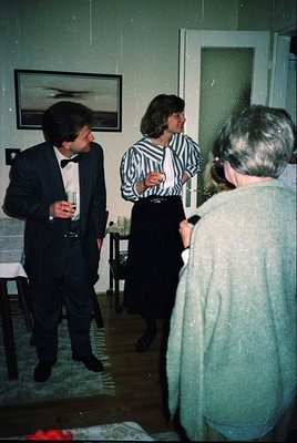Formal indoor gathering in a mid-century interior, featuring three adults in 1960s-70s attire: a man in a dark suit with a bo...