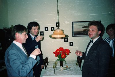 Four men in formal attire—dark suits, ties, and dress shirts—engage in conversation indoors, likely at a 1980s–1990s event. A...