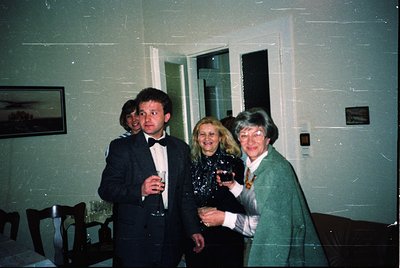 Three adults pose indoors, likely at a formal event in the 1990s. The man in a dark suit holds a wineglass; two women in form...