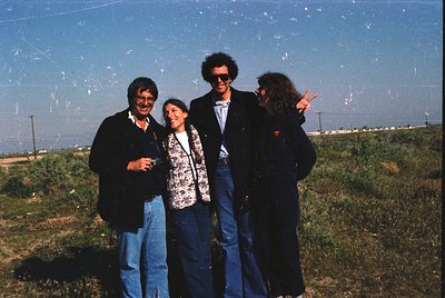 Four individuals pose outdoors in 1970s-style attire—men in blazers/sunglasses, women in patterned blouses and jeans—against ...