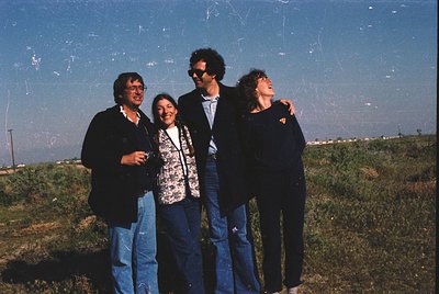 Four individuals pose outdoors in casual 1970s attire—men in blazers, women in patterned blouses and jeans—against a faded bl...