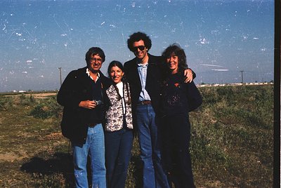 Four individuals pose outdoors in a grassy field under clear skies, likely late 20th century (1970s–1990s). The man on the le...
