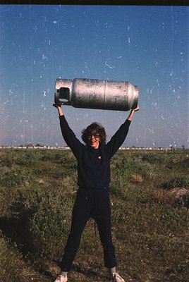 Person in outdoor setting holding large propane tank above head, likely mid-20th century. Clothing suggests casual, utilitari...